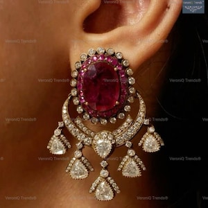 May include: Close-up of a gold-toned earring featuring a large, oval ruby surrounded by small, clear gemstones. The earring has a crescent-shaped design with dangling, pear-shaped and triangular gemstones. The earring is worn on an ear.