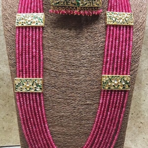 May include: A multi-strand pink beaded necklace with gold and green accents. The necklace has three rectangular pendants with floral designs and a matching pair of earrings with similar designs.