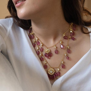 Può includere: Una collana dorata a più strati con perline di vetro rosa e dettagli floreali. La collana è indossata da una persona con i capelli scuri e una camicia bianca.