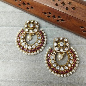May include: A pair of gold-toned earrings with a square top and a crescent-shaped bottom. The earrings feature clear and red gemstones, with a pearl drop and a border of small pearls. The earrings are on a gray surface.