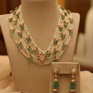Puede incluir: Un conjunto de collar y pendientes multicadena hecho de cuentas blancas, piedras verdes y detalles dorados. El collar tiene un diseño en capas con detalles rojos y dorados. Los pendientes tienen un diseño de borla con piedras verdes.