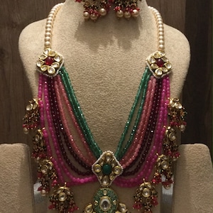 May include: A multi-strand necklace with pink, green, and maroon beads. The necklace has a large, ornate pendant with a green stone and gold accents. The necklace is paired with matching earrings with green stones and gold accents.