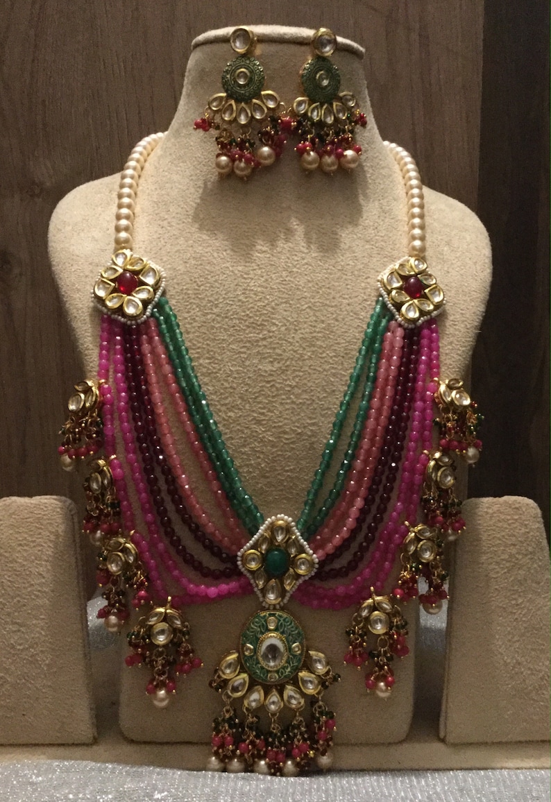 May include: A multi-strand necklace with pink, green, and maroon beads. The necklace has a large, ornate pendant with a green stone and gold accents. The necklace is paired with matching earrings with green stones and gold accents.