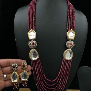 Puede incluir: Un collar de cuentas de color granate con detalles dorados y un colgante de espejo. El collar está formado por seis hileras de cuentas y tiene un juego de pendientes a juego.