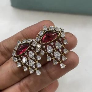 Può includere: Un paio di orecchini pendenti con una pietra rossa a forma di lacrima incastonata in una cornice di metallo dorato. Gli orecchini sono adornati con pietre di zircone cubico trasparenti che scendono a cascata sui lati.