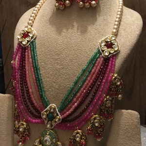 May include: A multi-strand necklace with pink, green, and burgundy beads, accented with gold and pearl embellishments. The necklace features a large, ornate pendant with a green stone and intricate details. Matching earrings with similar design elements are also included.