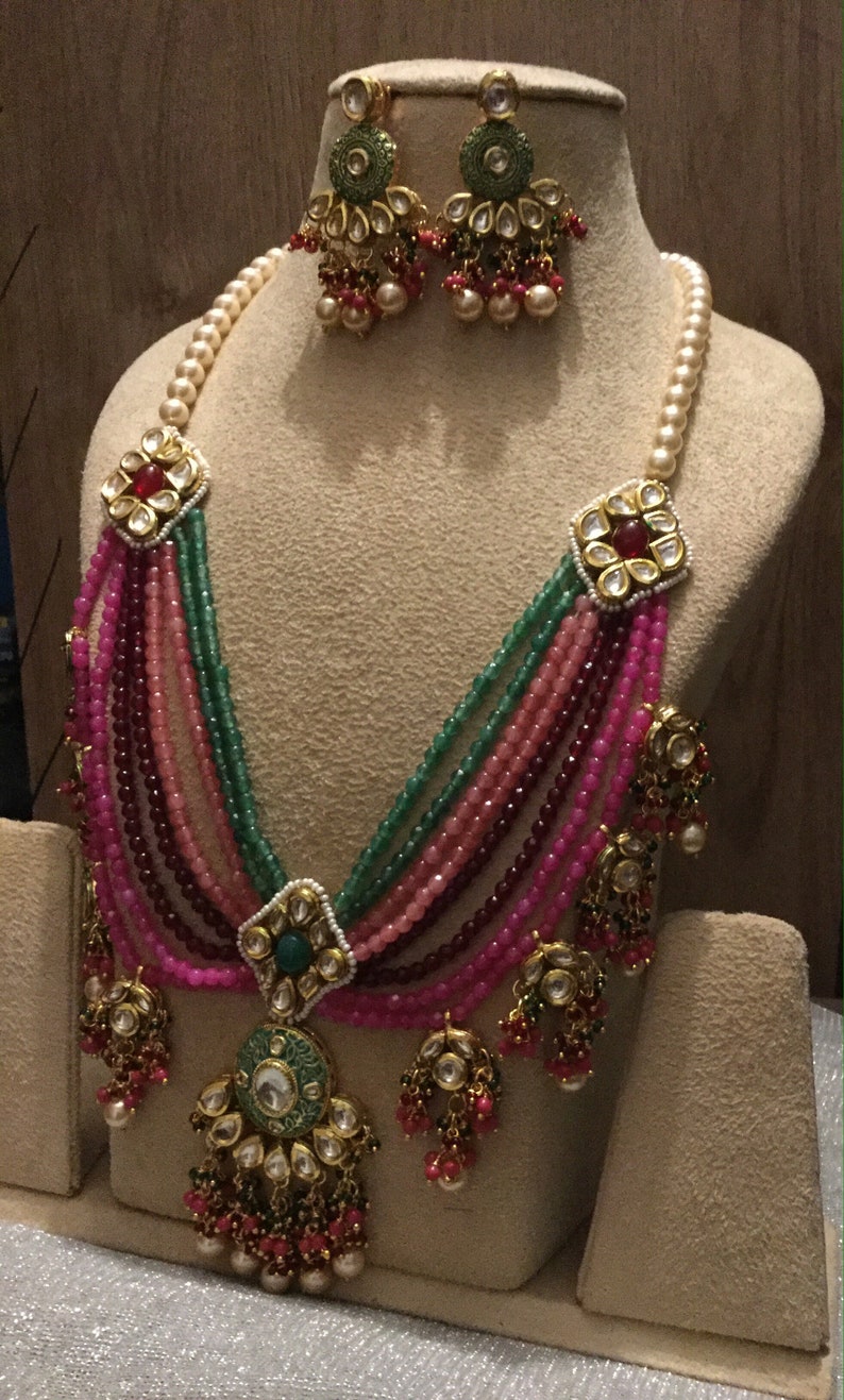 May include: A multi-strand necklace with pink, green, and burgundy beads, accented with gold and pearl embellishments. The necklace features a large, ornate pendant with a green stone and intricate details. Matching earrings with similar design elements are also included.