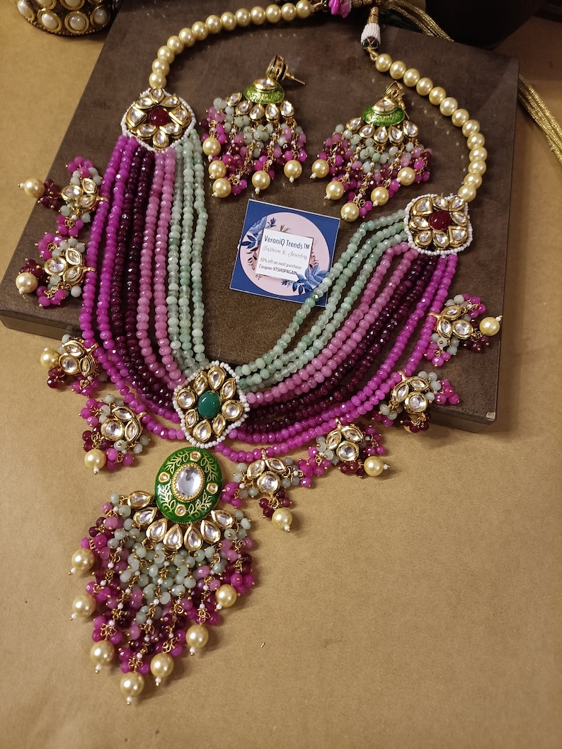 May include: A multi-strand necklace with pink, green, and white beads. The necklace has a large, ornate pendant with a green and gold design. The necklace is accompanied by a pair of matching earrings.