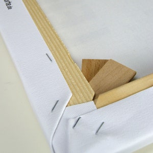 May include: Close-up of a white canvas stretched over a wooden frame. The canvas is secured with staples and wooden wedges are used to tighten the canvas.