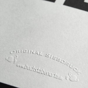May include: Close-up of a white, textured surface with the text "ORIGINAL SIEBDRUCK" and the website address "buchstabenorte.de" embossed in the surface.