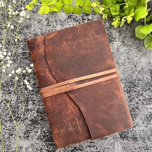 Könnte beinhalten: Ein braunes Lederjournal mit Wickelverschluss. Das Journal hat eine strukturierte Oberfläche und einen hellbraunen Lederriemen. Der Hintergrund ist eine strukturierte graue Oberfläche mit weißen Blumen und grünen Blättern.