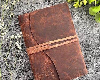 Könnte beinhalten: Ein braunes Lederjournal mit Wickelverschluss. Das Journal hat eine strukturierte Oberfläche und einen hellbraunen Lederriemen. Der Hintergrund ist eine strukturierte graue Oberfläche mit weißen Blumen und grünen Blättern.