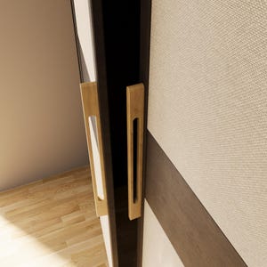 May include: Two wooden door handles with a rectangular design and a cutout for gripping. The handles are mounted on a dark-coloured door frame, with a light-coloured wall and a textured panel in the background. The wood has a natural finish.