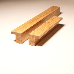 Puede incluir: Dos tiradores de armario de madera con diseño en forma de T. Los tiradores son de madera clara, posiblemente roble, y tienen un patrón de vetas natural. Uno de los tiradores es más grande que el otro. Los tiradores están diseñados para armarios o cajones.