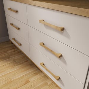 May include: A white six-drawer dresser with light wood trim and handles. The wooden handles are a light brown color and have a unique, curved design. The dresser sits on a light wood floor.