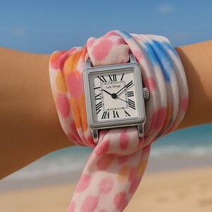 May include: A silver Lady Mirage watch with a square face and Roman numerals is wrapped in a colourful scarf. The scarf features pink, orange, and blue tie-dye patterns. The watch is on a wrist.