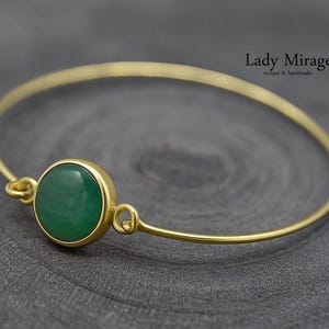 Könnte beinhalten: Ein goldfarbenes Armreif-Armband mit einem runden, grünen Edelstein, der in einer goldenen Lünette gefasst ist. Das Armband hat ein schlichtes, elegantes Design und liegt auf einer dunkelgrauen Oberfläche. Der Text "Lady Mirage" ist sichtbar.