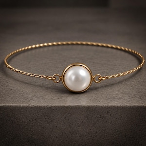 May include: A gold-toned bracelet featuring a thin, twisted metal band and a single, centred, round white pearl set in a smooth gold-toned bezel. The pearl is connected to the twisted band by two small circular metal loops on either side.