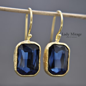 Pode incluir: Um par de brincos de ouro com pedras retangulares azul-escuras. Os brincos têm um design simples e elegante.