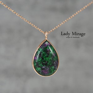 Op de afbeelding: Een ketting met een traanvormige hanger, bezet met een groene en paarse edelsteen in een gouden zetting. De ketting is van goud en heeft een delicaat schakelschema. De hanger is gemarkeerd met "Lady Mirage unique & handmade".