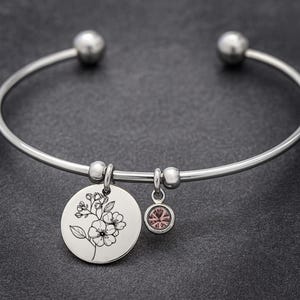 Könnte beinhalten: Ein silberfarbenes Armreif-Armband mit einem Blumenanhänger und einem kleinen rosa Edelstein-Charm. Der runde Anhänger zeigt ein detailliertes schwarz-weißes Blumendesign. Das Armband hat ein schlichtes, elegantes Design.