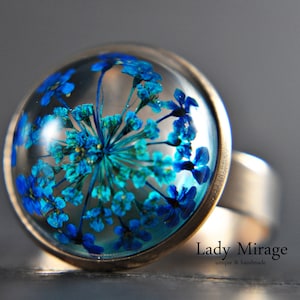 Puede incluir: Un anillo de plata con una flor azul y turquesa prensada dentro de una cúpula de vidrio transparente. El anillo está colocado sobre una superficie gris. El texto "Lady Mirage unique & handmade" es visible debajo del anillo.