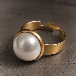 Op de afbeelding: Een goudkleurige ring met een grote, gladde, witte bolvormige parel in een ronde zetting. De ringband is breed en heeft een open, verstelbaar ontwerp aan de achterkant, afgewerkt met een matte metallic uitstraling over de gehele structuur.