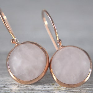 Könnte beinhalten: Ein Paar roségoldene Ohrhänger. Jeder Ohrring hat einen runden, hellrosa Stein, der in einen roségoldenen Rahmen gefasst ist. Die Ohrringe haben ein schlichtes, elegantes Design mit einem gebogenen Haken.