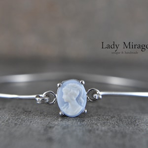 Könnte beinhalten: Ein silbernes Armreif-Armband mit einem weißen Kamee, das das Profil einer Frau zeigt. Das Kamee ist in einem silbernen Rahmen gefasst und mit einem silbernen Verschluss am Armband befestigt. Das Armband befindet sich auf einem grauen Hintergrund. Lady Mirage einzigartig & handgefertigt.