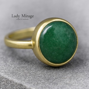 Könnte beinhalten: Ein goldfarbener Ring mit einem großen, runden, polierten grünen Stein. Der Ring hat eine einfache Lünettenfassung, und der Ring ist glatt und abgerundet. Der Text "Lady Mirage unique & handmade" ist oben links zu sehen.