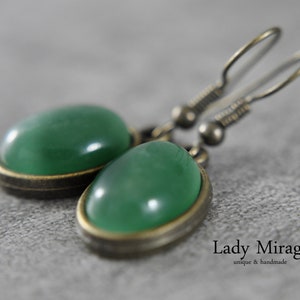 Op de afbeelding: Een paar groene edelsteen oorbellen met een messing setting en zilveren haken. De oorbellen zijn ovaalvormig en hebben een gladde, gepolijste afwerking.  "Lady Mirage" staat in zwarte tekst op een witte achtergrond onder de oorbellen.