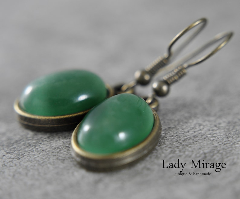 Op de afbeelding: Een paar groene edelsteen oorbellen met een messing setting en zilveren haken. De oorbellen zijn ovaalvormig en hebben een gladde, gepolijste afwerking.  "Lady Mirage" staat in zwarte tekst op een witte achtergrond onder de oorbellen.