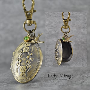 Può includere: Un ciondolo ovale dorato con un design floreale e una piccola pietra preziosa verde. Un piccolo ciondolo a forma di uccello pende dal ciondolo. Il ciondolo è attaccato a una catena dorata con un moschettone.