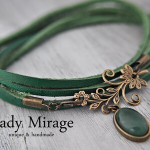Op de afbeelding: Een groene leren wikkelarmband met een bronzen bloemenhanger en een groene edelsteen. De armband heeft een sluiting en de hanger heeft een delicaat bloemenmotief.