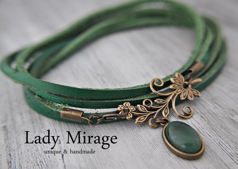 Op de afbeelding: Een groene leren wikkelarmband met een bronzen bloemenhanger en een groene edelsteen. De armband heeft een sluiting en de hanger heeft een delicaat bloemenmotief.