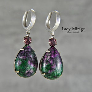 Peut inclure: Une paire de boucles d'oreilles créoles en argent avec une pierre en forme de larme verte et violette et un petit accent de pierre rose. Les boucles d'oreilles sont étiquetées "Lady Mirage unique & handmade".