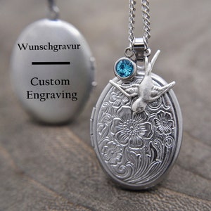 Könnte beinhalten: Silberne ovale Halskette mit einem blauen Edelstein und einem Vogel-Anhänger. Das Medaillon hat ein gravierter Blumenmuster und die Rückseite ist leer für eine individuelle Gravur. Der Text "Wunschgravur" und "Custom Engraving" ist auf der Rückseite des Medaillons sichtbar.