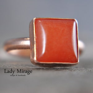 Puede incluir: Un anillo de forma cuadrada con una piedra preciosa naranja brillante engastada en una banda de oro rosa.