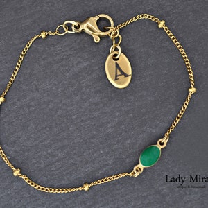 Könnte beinhalten: Goldkette Armband mit einem grünen ovalen Stein und einem Anhänger mit dem Buchstaben A. Das Armband wird von Lady Mirage hergestellt, einem Unternehmen, das sich auf einzigartige und handgefertigte Schmuckstücke spezialisiert hat.