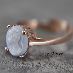 Può includere: Un delicato anello con un cammeo che presenta il profilo di una donna in bianco su uno sfondo blu chiaro. L'anello è in oro rosa e ha una fascia semplice.