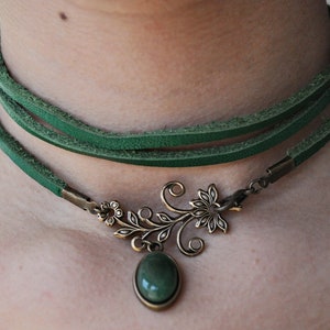 Op de afbeelding: Een groene leren choker ketting met een bronzen hanger met een gestileerd bloemmotief en een groene edelsteen.