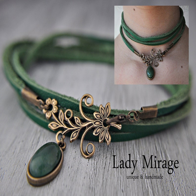 Op de afbeelding: Een groene leren choker ketting met een bronzen bloemenhanger en een groene edelsteen. De ketting is gelaagd en heeft een sluiting. De hanger heeft een delicaat bloemenontwerp met een enkele groene edelsteen in het midden. De ketting is handgemaakt en uniek.