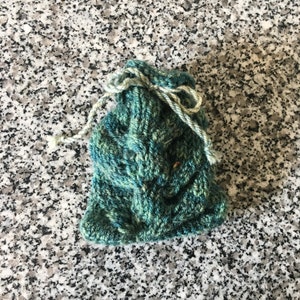 May include: A small, hand-knitted bag in shades of teal and green, with a drawstring closure. The bag is made of textured yarn, and the drawstring is a lighter shade of green. The bag is likely used for small items or gifts.