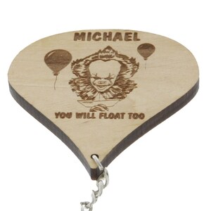 Könnte beinhalten: Holzschlüsselanhänger in Form eines Heißluftballons mit einer Zeichnung von Pennywise dem Clown aus dem Film "Es" und dem Text "YOU WILL FLOAT TOO". Der Schlüsselanhänger trägt auch den Namen "MICHAEL" oben und eine silberne Kette.