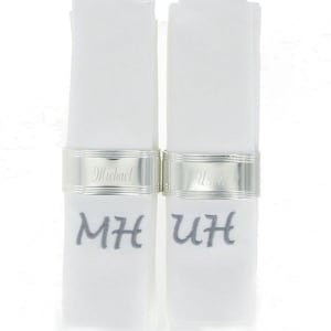 May include: Two white linen napkins with silver napkin rings. The rings are engraved with the names "Michael" and "Ursula". The napkins are embroidered with the initials "MH" and "UH" in grey thread.