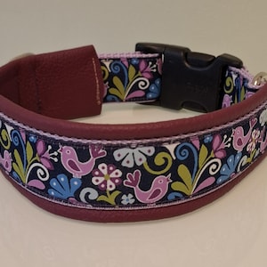 May include: A dog collar with a burgundy leather exterior and a patterned fabric interior. The fabric features a navy blue background with pink birds, blue flowers, and green and yellow foliage. A black plastic buckle secures the collar.
