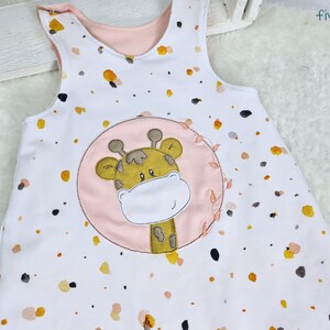 May include: A white sleeveless onesie with a pink and yellow giraffe applique. The onesie has a white background with small yellow and pink spots.