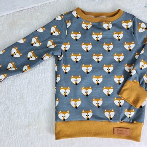 Könnte beinhalten: Ein blauer Langarm-Sweatshirt mit einem Fuchsgesichtsmuster. Der Sweatshirt hat einen senfgelben Besatz an Hals, Ärmelbündchen und Saum. Das Markenetikett "fivekidz" ist auf der rechten unteren Seite des Sweatshirts sichtbar.
