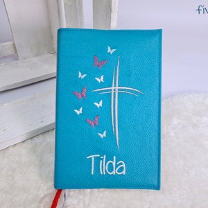 Puede incluir: Un diario de cuero azul turquesa con una cruz blanca y mariposas rosas bordadas en la parte delantera. El nombre "Tilda" est&aacute; bordado debajo de la cruz.