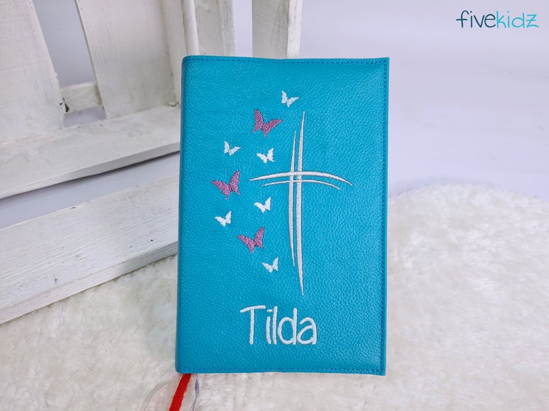 Puede incluir: Un diario de cuero azul turquesa con una cruz blanca y mariposas rosas bordadas en la parte delantera. El nombre "Tilda" est&aacute; bordado debajo de la cruz.